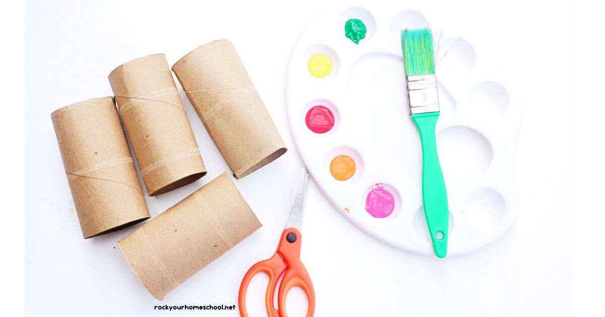 Four toilet paper rolls, scissors, white paint palette, green paint brush, and paint.