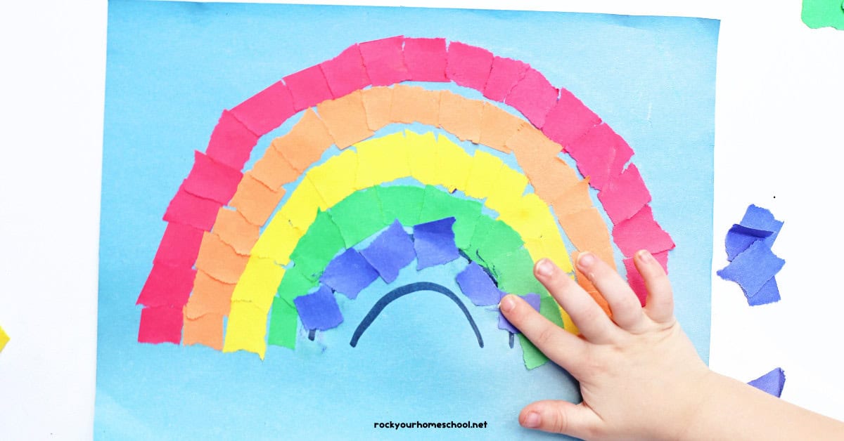 Child glueing piece of blue torn paper on rainbow craft.