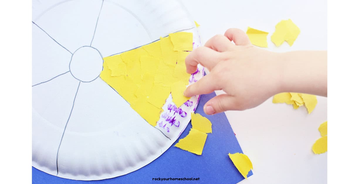 Child using glue stick to add yellow torn paper squares to beach ball paper plate craft.