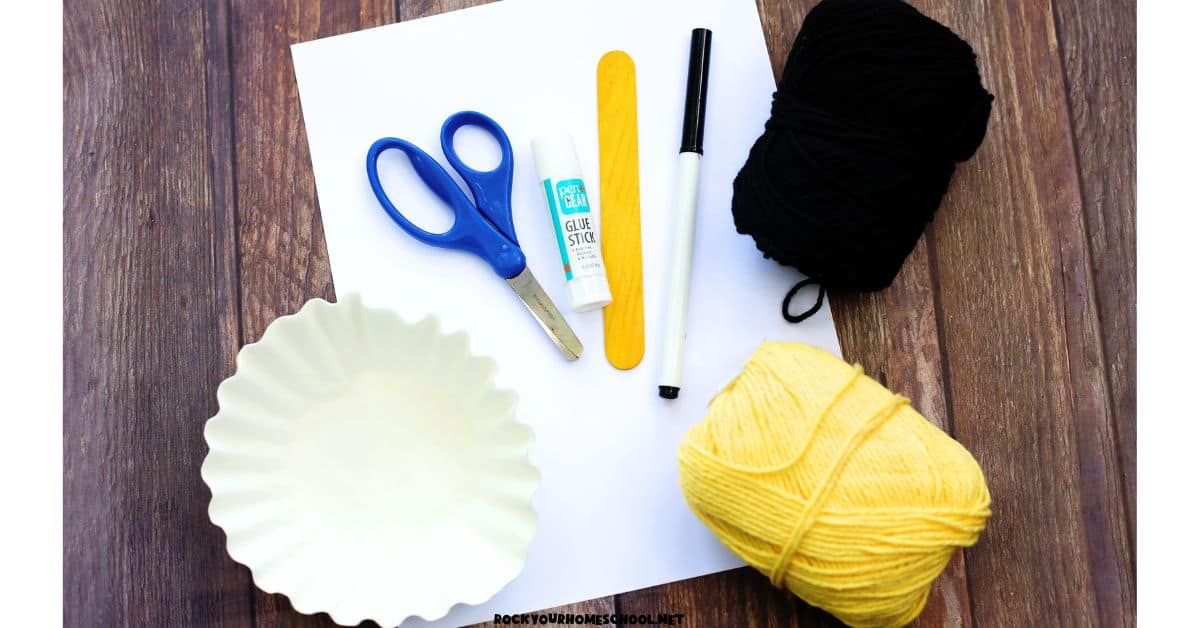 Supplies for Little Red Riding Hood craft with white paper, scissors, glue stick, yellow wood craft stick, black marker, black and yellow yarn, and coffee filters.