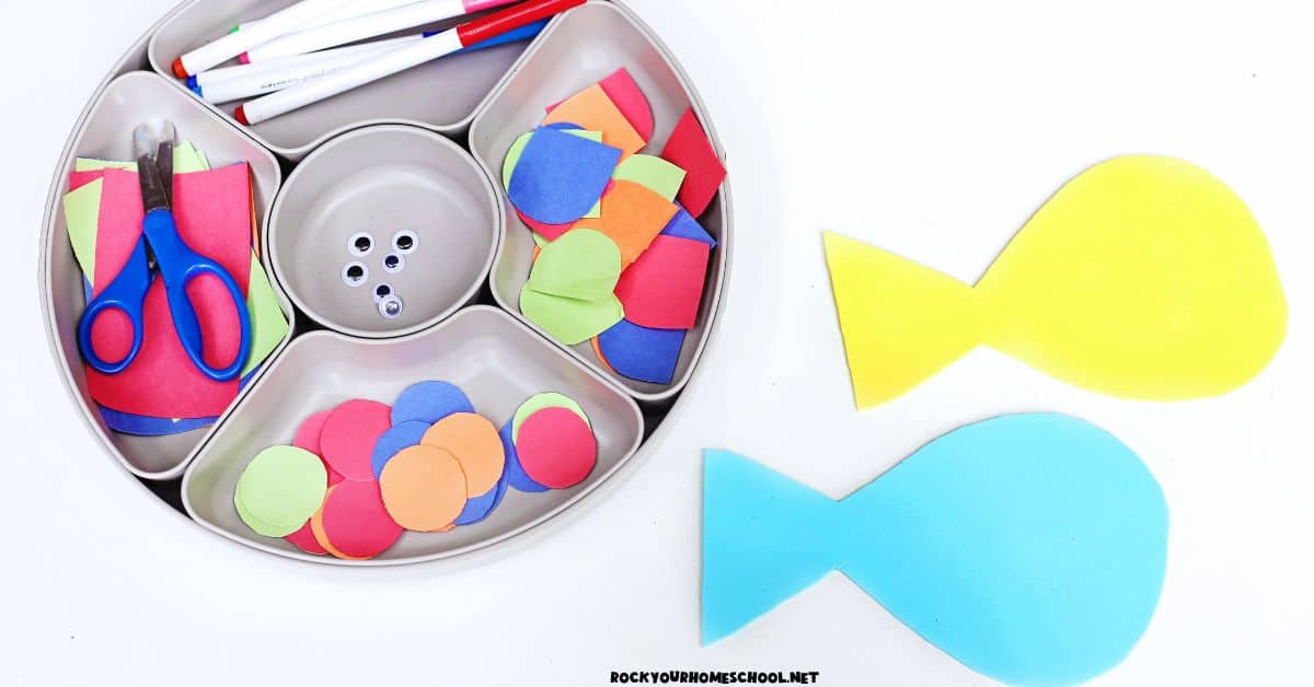 Activity tray with markers, googly eyes, scissors, colorful paper in different shapes, and yellow and light blue paper fish.