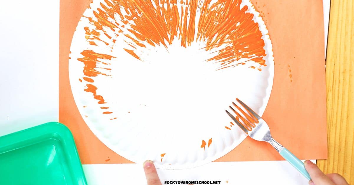 Child using fork with orange paint on paper plate.