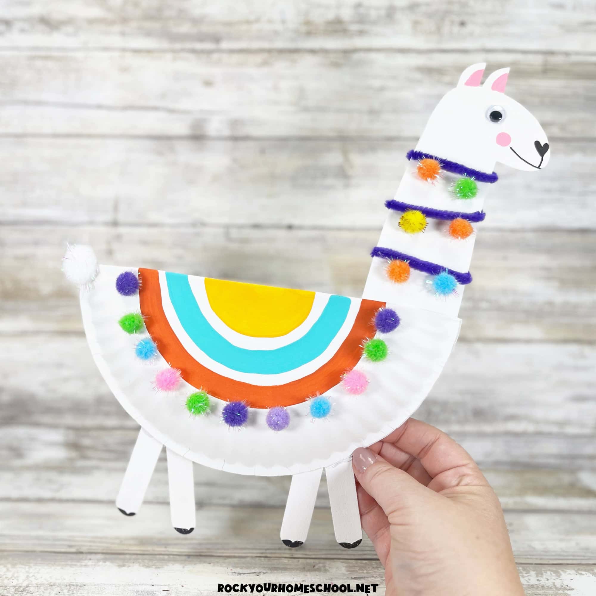 Woman holding example of paper plate llama craft for kids with pom poms.