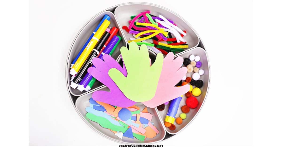 Example of easy monster handprint activity tray with construction paper handprints, markers, pipe cleaner pieces, pom poms, glue stick, and paper shapes.