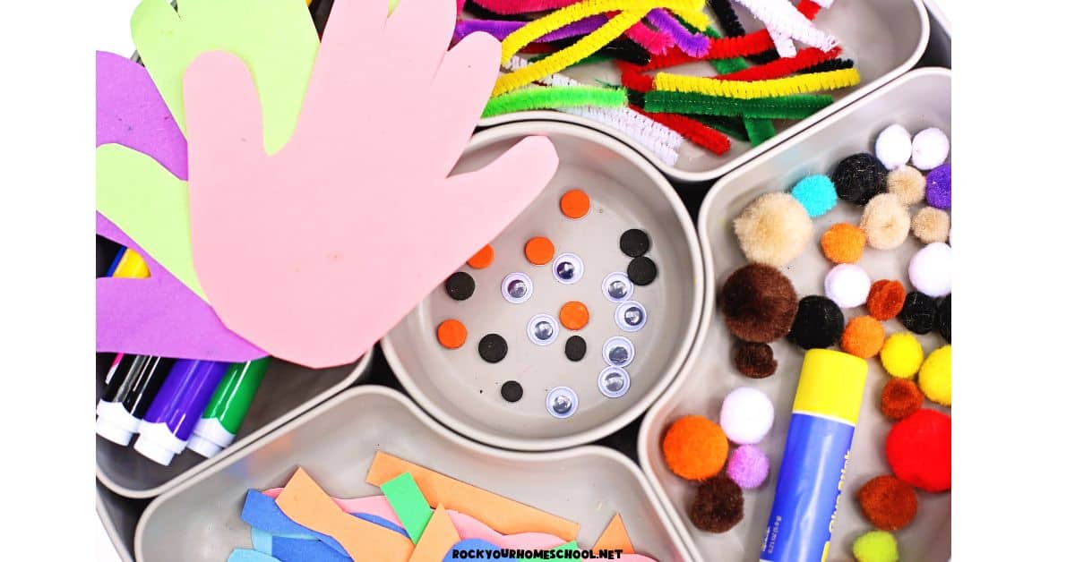 Construction paper handprints, googly eyes, pom poms, glue stick, paper shapes, markers, and pipe cleaners on activity tray.