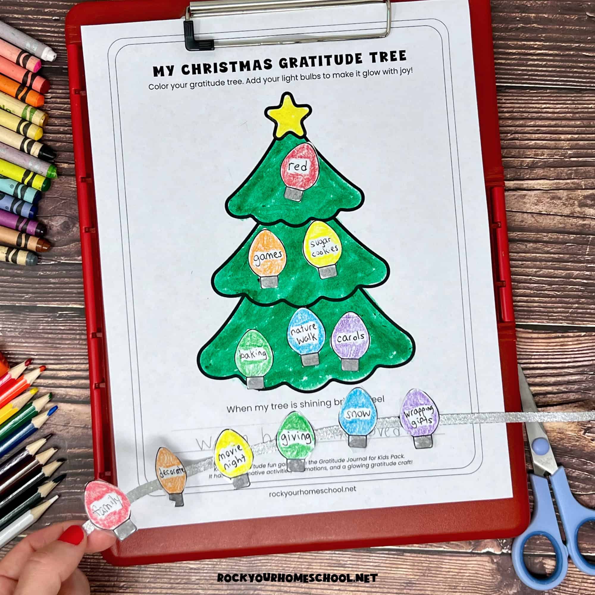 Woman holding red clipboard with free printable Christmas gratitude tree activity and Christmas lights on silver ribbon.