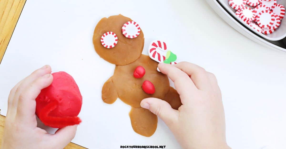 Child adding red playdough balls to gingerbread playdough character with peppermint embellishments.
