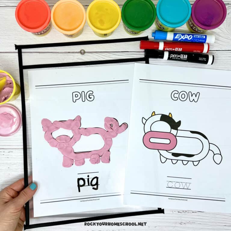 Woman holding printable pig playdough mat with cow playdough mat in background with rainbow of playdough cans and dry erase markers.