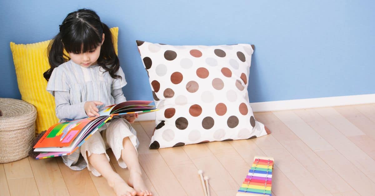 Young girl looking at book in corner of room with pillows.