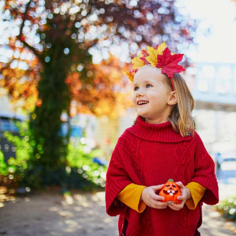 Preschool girl holding pumpkin and wearing autumn leaves headband while looking at a tree to feature these fun preschool fall themes.